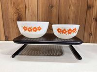 Two white milk glass mixing bowls with orange floral decoration, displayed side by side on a black metal perforated stand, showing size difference