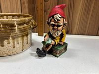 Glazed pottery bowl in beige and brown drip glaze with two handles next to ceramic gnome figurine wearing red hat sitting on green book