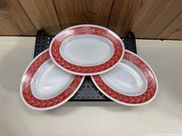 Three white oval platter plates with red laurel leaf patterned band around the rim placed on a black perforated platform.