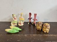 Photo showing four sets of vintage ceramic salt and pepper shakers on a dark wooden surface, including corn cob, musical couple, spoon and fork, and dog figurines.