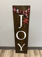 Full view of the wooden porch sign with JOY in vertical white letters, stained wood, plaid fabric bow, and red star ornament.