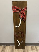 Vertical wooden porch sign with brown stain, white letters 'J' and 'Y', red and green plaid bow tied at top, and grapevine wreath with red berries as the letter 'O'.