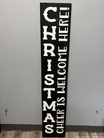 Full height view of the wooden Christmas porch sign showing text 'CHRISTMAS CHEER IS WELCOME HERE!' vertically arranged in white lettering on black wood.