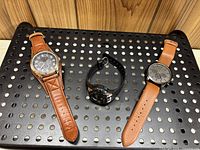 Photo of three wristwatches displayed on a black perforated metal surface against a wood background. Two watches have brown leather straps, one has black rubber strap. Shows overall condition and details of each watch.