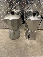 Two Bialetti aluminum espresso makers side by side on a metallic surface showing overall shape and condition.