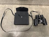 Black Bushnell Insta-Focus Zoom binoculars next to accompanying black carrying case with strap on metal surface.