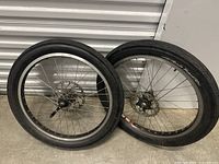Two cruiser bike wheels with metal spokes and disc brake mounts leaning against a corrugated metal wall.