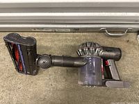 Dyson handheld vacuum cleaner body and detachable cleaning head shown assembled on a concrete floor beneath a metal surface.