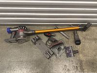 Photo showing the Dyson vacuum cleaner main body, long extension tube, charging cable, and various tool attachments laid out on a concrete floor.
