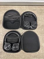 Two black over-ear headphones, each placed inside a black semi-hard carrying case on a concrete floor, cables included.