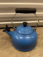 Front-side view of blue Le Creuset tea kettle, showing round body, lid, handle, and spout with lever