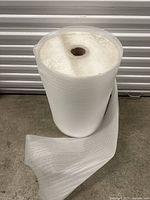 Single large roll of white foam packing material with textured surface and hollow core shown standing upright on concrete floor in front of a metal shutter door.