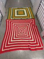 Two vintage crocheted blankets laid flat on a floor in a hallway. One blanket is red and cream with concentric square stripes, the other is green, yellow, and cream with square stripe pattern.