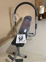 Front view of Ab King Pro exercise bench with user manual placed on the seat, showing padded backrest partially covered in plastic and foam grips on handles and footrest.