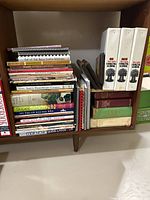 Books and wooden baking plaques in a wooden shelf. Visible book titles include 'Out of Old Nova Scotia Kitchens','Campbell's 100 Best Recipes', 'Simply HeartSmart Cooking', 'Super Salads', 'Light & Easy Chinese', 'Land O Lakes Classic Cookies','Five Seasons Cranberry Book', 'Better Homes and Gardens Home Canning Cook Book', and more.