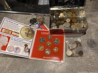 Overview of lot showing Royal Mint coin jacket with informational panels, small glass jar, plastic bag, and decorative tin box of coins