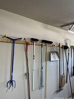 Full view of tools hanging on wooden rack in garage showing blue cultivator, orange lopper, metal rake and brooms