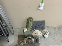 Overall group of garden decor items arranged on floor against wall, showing sleeping angel, cat, rain gauge, bird, sundial, fairy sign.