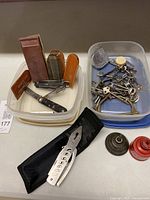 Photo showing various multi-tools, whetstones, and a collection of keys in plastic containers, along with two small vintage oil cans on the side