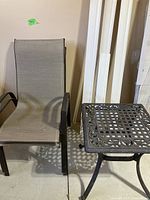 Photo of one brown metal strap chair with armrests and taupe/grey fabric seat next to black metal side table with lattice top and decorative scrollwork.