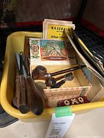 First image of yellow container holding assorted wooden pipes, pipe cutters, a cigar box with tobacco pipes inside, vintage Kellogg's baseball booklets, and other small vintage items.