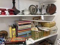 Shelf holding a variety of vintage and assorted sewing pattern books and hardcover sewing books, silver serving pieces on upper shelf