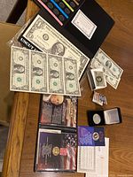 Image shows the full collection laid out on a wooden surface including uncut sheet of $1 bills, John F. Kennedy half dollar in flip case, John Adams Presidential Dollar with stamp, and paper currency albums.