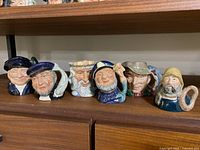 Six Toby jugs displayed on a wooden shelf, showing the full assortment including characters and handle shapes
