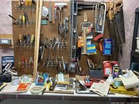 Wide view of pegboard and workbench loaded with hand tools and hardware, including saws, screwdrivers, pliers, clamps, stapler, and packaging for staples and zip ties.