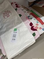 Stack of folded unfinished quilts showing the variety of patterns including embroidery, applique, and patchwork in red, pink, green, white, and cream fabrics.