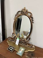 Vintage oval mirror with ornate scroll detail and attached matching wooden shelf with gold finish. Two small framed pictures placed in front.