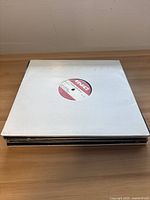 Stack of 10 vinyl records in paper sleeves stacked horizontally on wooden surface.