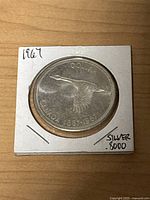 Front side of the 1967 Canadian silver loonie coin showing a flying Canada goose and inscriptions 'DOLLAR CANADA 1867-1967', with silver .8000 indication on paper holder.