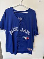 Front view of Blue Jays baseball jersey, showing team name and logo on chest area, button closure, and Majestic patch on bottom hem.