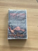 Front of sealed cassette tape with album artwork showing title and artist credits.