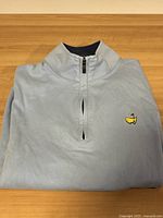 Folded light blue long sleeve quarter zip pullover with zippered collar detail and Masters logo embroidered on chest.