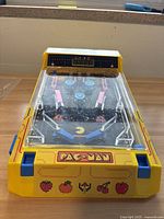 Front top-down view of miniature yellow Pac-Man arcade game showing detailed pinball playfield with Pac-Man and ghosts inside clear plastic cover and score display on yellow casing with fruit graphics.