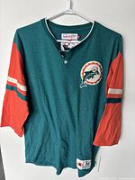 Front view of Mitchell Ness Throwbacks Miami Dolphins shirt, teal with orange sleeves and logo patch on chest.