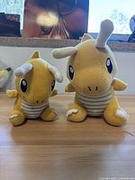 Front view of two Pokemon Dragonite plush toys, one larger and one smaller, placed on a wooden surface next to a window.