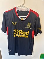 Front view of the Rangers navy blue 150 year edition football jersey with gold sponsor logos and red shoulder accents.