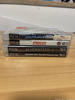 Four sealed PS3 games stacked to show spine titles: Metal Gear Rising, Syndicate, Resident Evil 6, Resident Evil Operation Raccoon City.