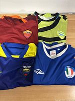 Photo of four soccer jerseys from Italy, Ecuador, Arsenal, and Roma folded and laid on table.