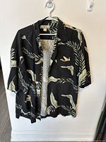 Front view of black Tommy Bahama short sleeve silk shirt with cream leaf and bird pattern hanging on a white wall.
