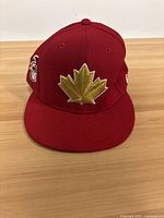 Front view of red New Era 9Twenty baseball cap with gold maple leaf embroidered on front.