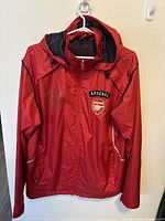 Photo of a red Arsenal FC training jacket with hood hung on a plastic white hanger showing front view with Arsenal crest on chest.