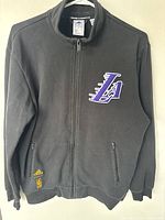 Full front view of black LA Lakers zip-up sweater showing logos and zipper pockets