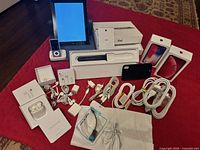 Apple electronics and accessories arranged on floor, including iPad 3G 64GB with box and a black case, two iPhone boxes with packaging, iPod Nano, power cables, adapters, AirPods boxes, and various cords and chargers.