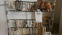 Two rows of assorted bracelets hanging on metal bars showing white beaded, wood and black/gold beaded bracelets on upper row and assorted charm and linked bracelets on lower row.