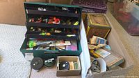 Overview photo showing fishing tackle box filled with colorful freshwater lures and flies, three fly fishing reels placed separately, yellow vintage metal bait canteen, and various small accessory packets and boxes.