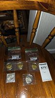 Coins seen on wooden chair, showing $5 and $10 commemorative coins in plastic sleeves and bags.